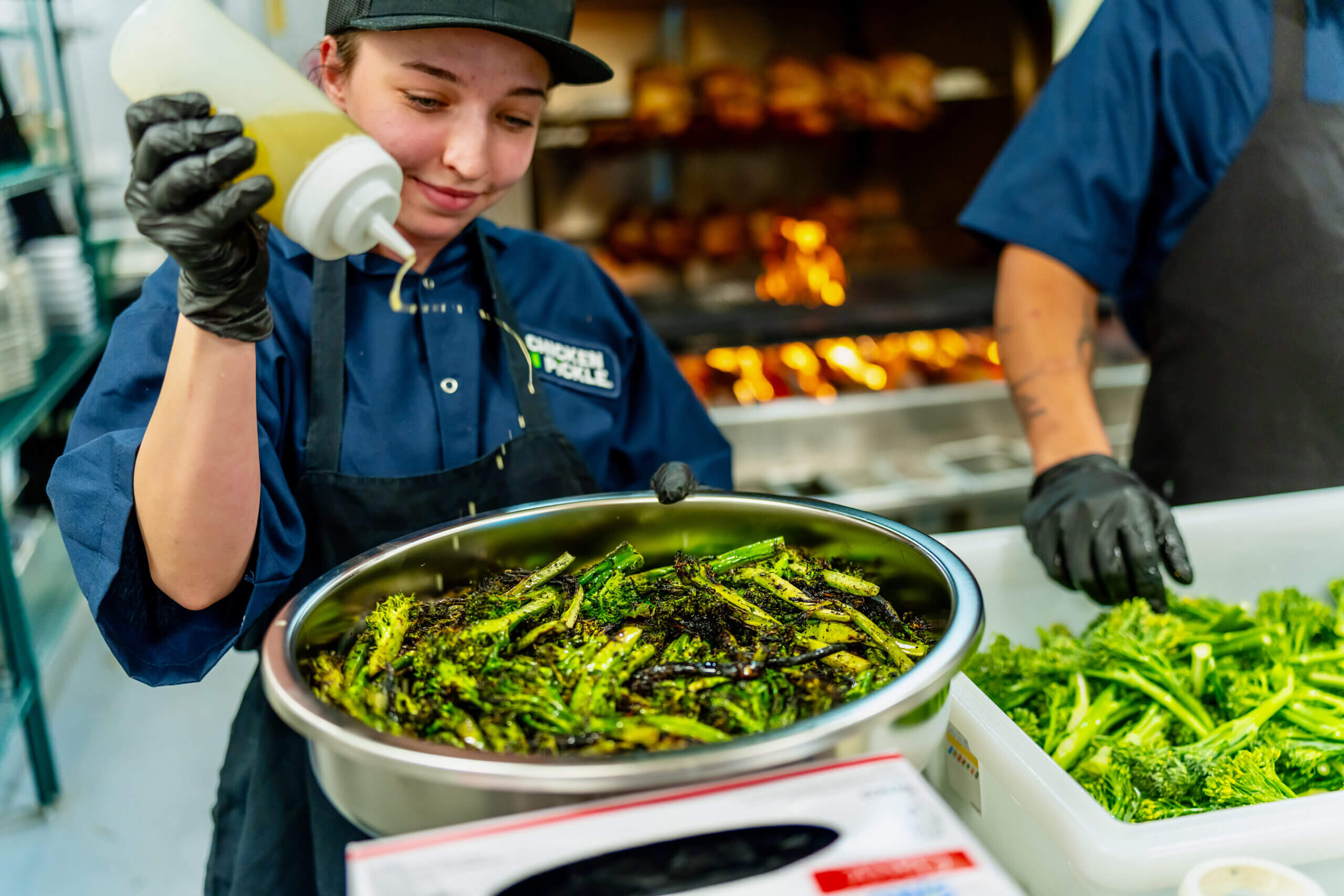 A chef wearing a blue uniform and black gloves pours oil over a large bowl of cooked broccolini, with fresh broccolini and a grill fire visible in the background.