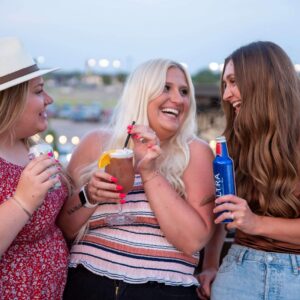 Three women are smiling and laughing together outdoors at dusk, each holding a drink. They appear to be enjoying a casual, cheerful moment, with lights and greenery blurred in the background.