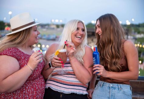 Three women are smiling and laughing together outdoors at dusk, each holding a drink. They appear to be enjoying a casual, cheerful moment, with lights and greenery blurred in the background.