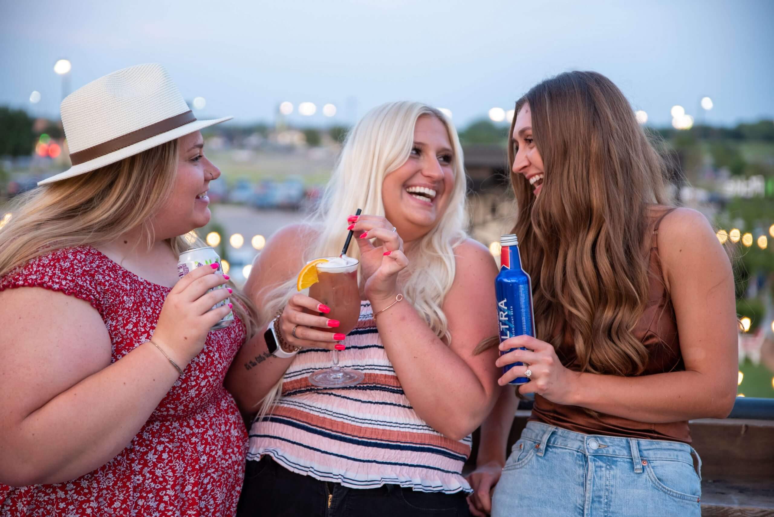 Three women are smiling and laughing together outdoors at dusk, each holding a drink. They appear to be enjoying a casual, cheerful moment, with lights and greenery blurred in the background.
