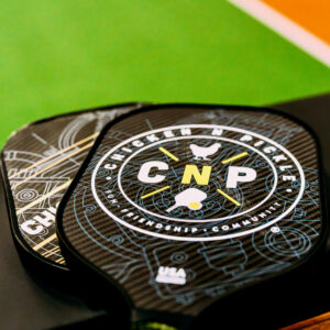 A close-up of two pickleball paddles with a “Chicken N Pickle” logo, resting on a black bench next to a green and orange pickleball court.