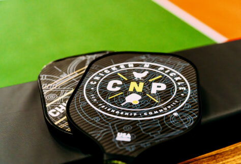 A close-up of two pickleball paddles with a “Chicken N Pickle” logo, resting on a black bench next to a green and orange pickleball court.