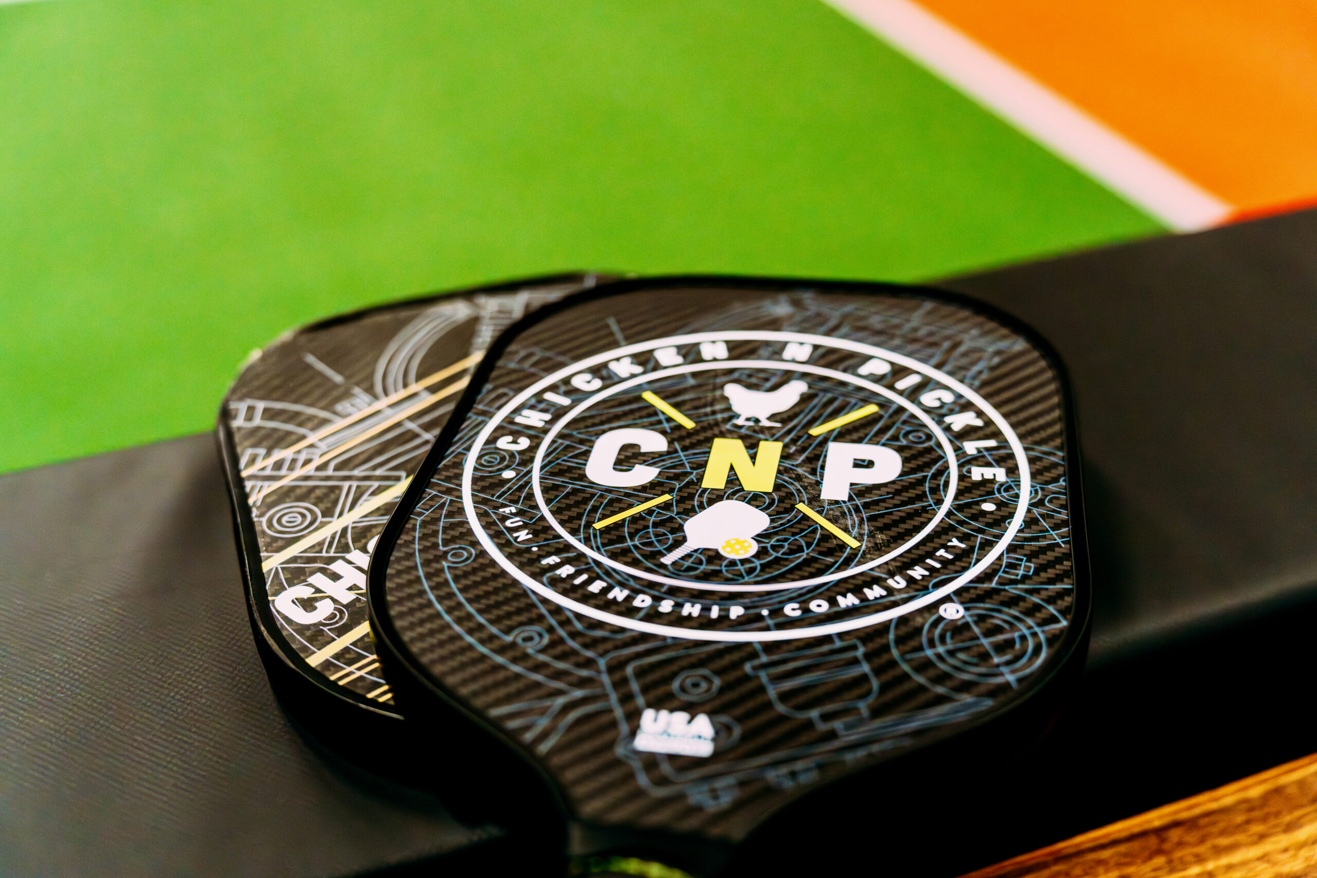 A close-up of two pickleball paddles with a “Chicken N Pickle” logo, resting on a black bench next to a green and orange pickleball court.