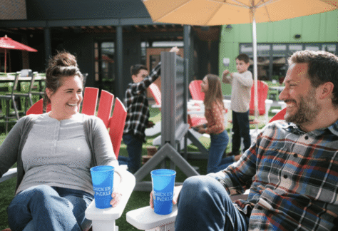 A woman and a man sit smiling on red chairs outdoors, holding blue cups. In the background, children play a large Connect Four game on a grassy area under umbrellas and the sun.
