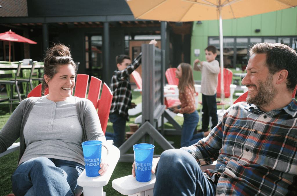 A woman and a man sit smiling on red chairs outdoors, holding blue cups. In the background, children play a large Connect Four game on a grassy area under umbrellas and the sun.
