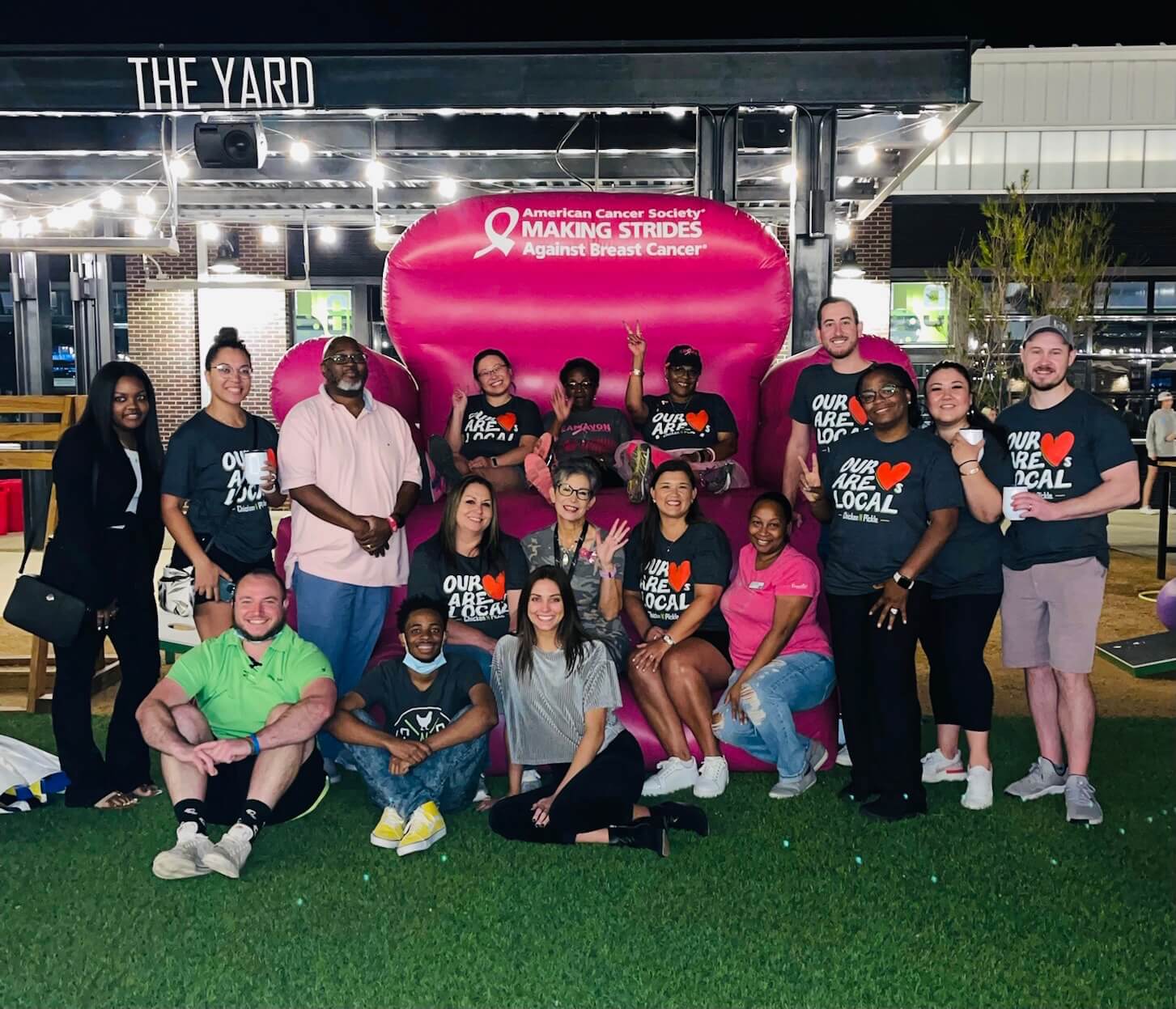 A diverse group of people pose together at night in front of a large pink chair and a sign for the American Cancer Society’s Making Strides Against Breast Cancer event. Many wear shirts with “Our Heart is Local” printed on them.