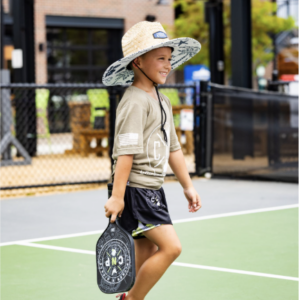A young boy in a wide-brimmed hat, t-shirt, and shorts walks on a pickleball court at Doggy Days at Chicken N Pickle Allen, smiling and holding a pickleball paddle. An outdoor sports facility is visible in the background.
