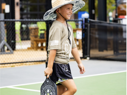 A young boy in a wide-brimmed hat, t-shirt, and shorts walks on a pickleball court at Doggy Days at Chicken N Pickle Allen, smiling and holding a pickleball paddle. An outdoor sports facility is visible in the background.