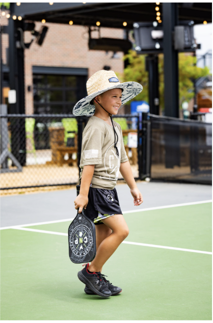 A young boy in a wide-brimmed hat, t-shirt, and shorts walks on a pickleball court at Doggy Days at Chicken N Pickle Allen, smiling and holding a pickleball paddle. An outdoor sports facility is visible in the background.