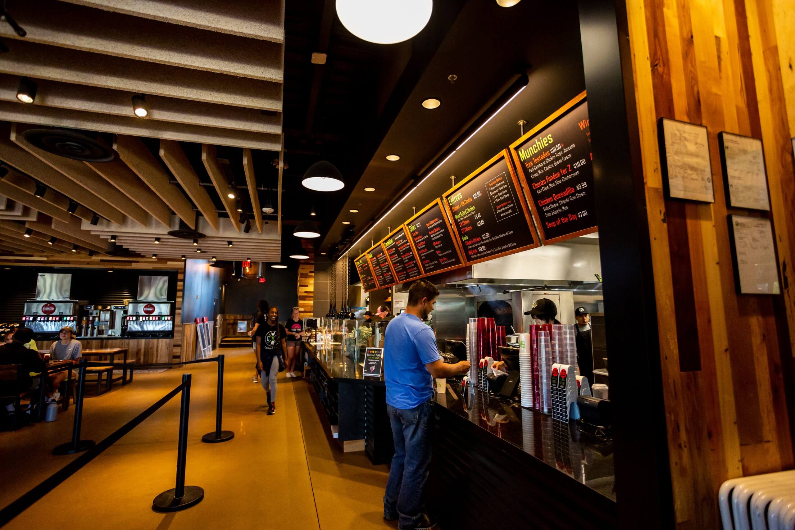 A man stands at the counter of a casual restaurant, looking at the menu boards overhead while staff prepare food behind the counter. The interior features warm lighting, wood accents, and several people dining.