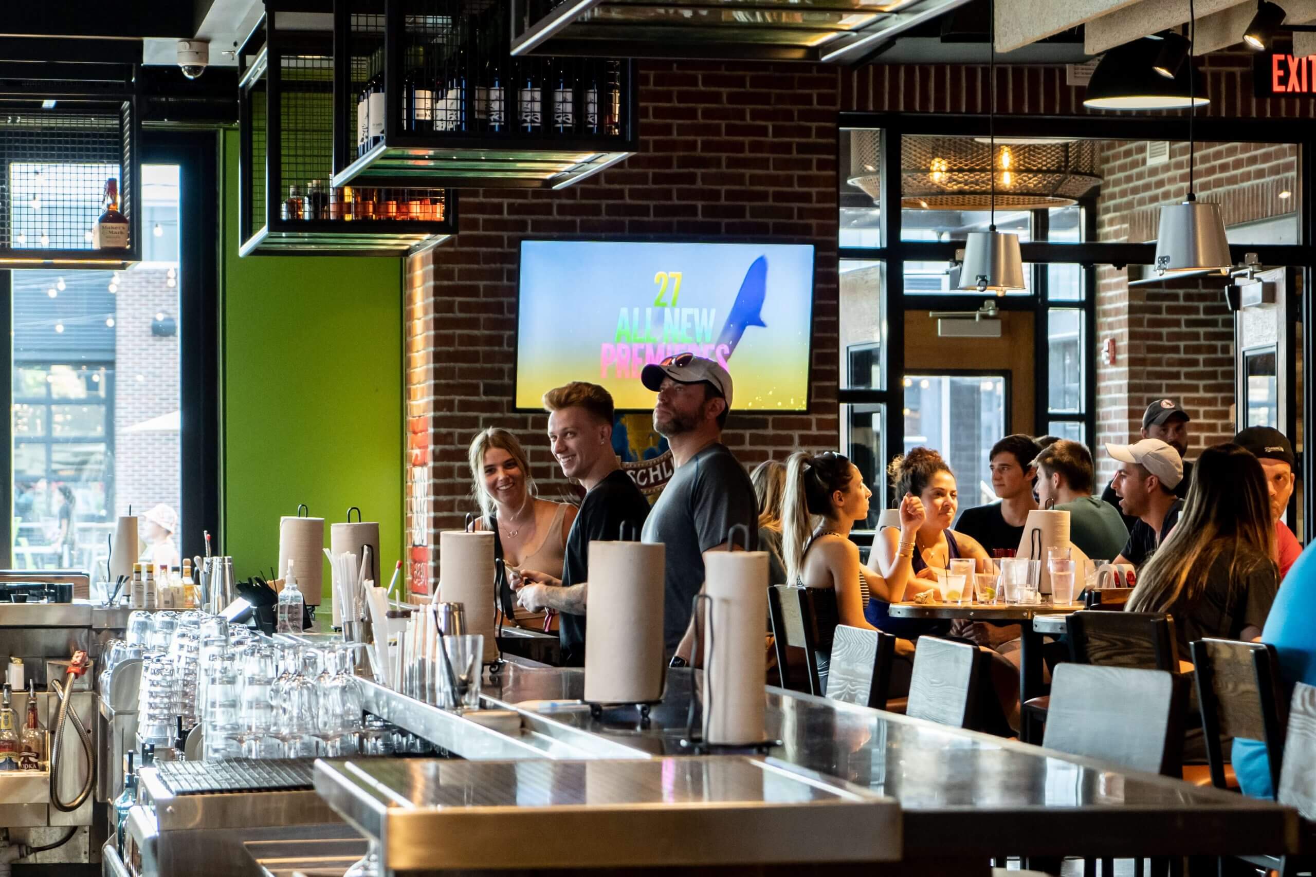 People gather and socialize inside a lively bar or restaurant with brick walls. Some stand at the bar, while others sit at tables. Glasses and bottles are visible, and a TV screen is displayed in the background.