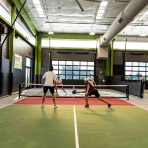 Four people play a doubles pickleball match indoors on a green and red court, with two players at the net and two near the baseline. Large windows and ceiling fans are visible in the background.