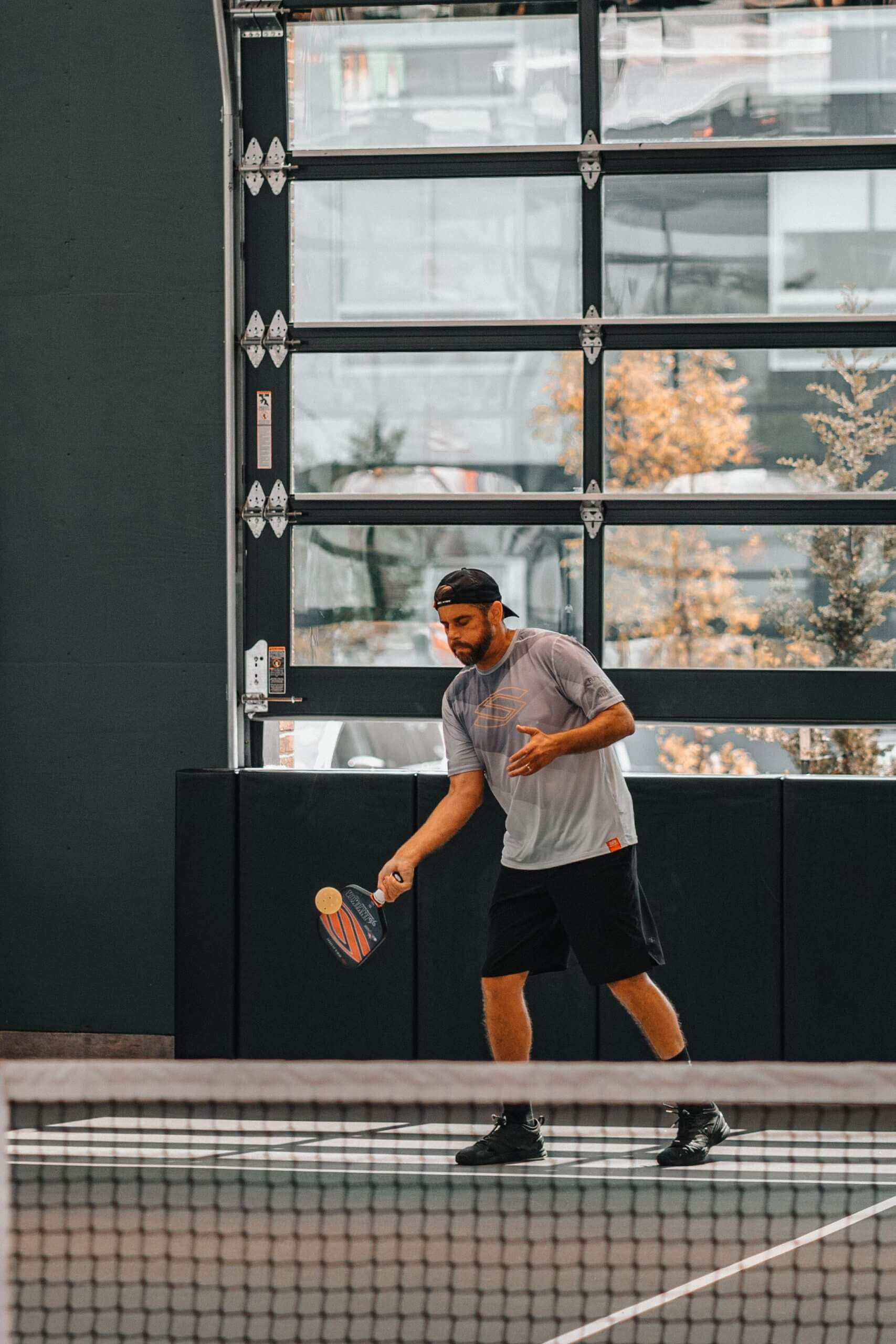 A man in sportswear prepares to hit a ball with a paddle on an indoor court. He stands behind the net, focused, with a garage-style door and trees visible in the background.