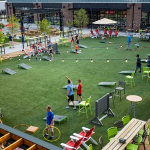 People play lawn games like cornhole and Connect Four on a large artificial grass yard surrounded by tables and chairs, with string lights overhead and a brick building in the background.