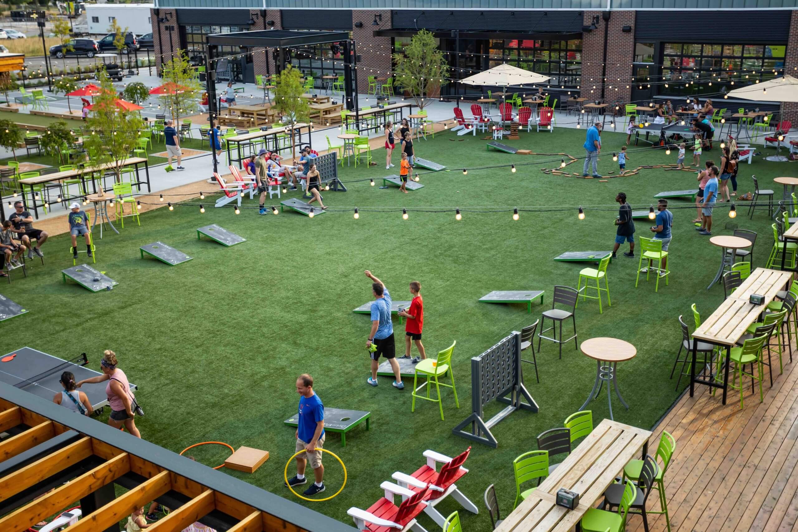 People play lawn games like cornhole and Connect Four on a large artificial grass yard surrounded by tables and chairs, with string lights overhead and a brick building in the background.