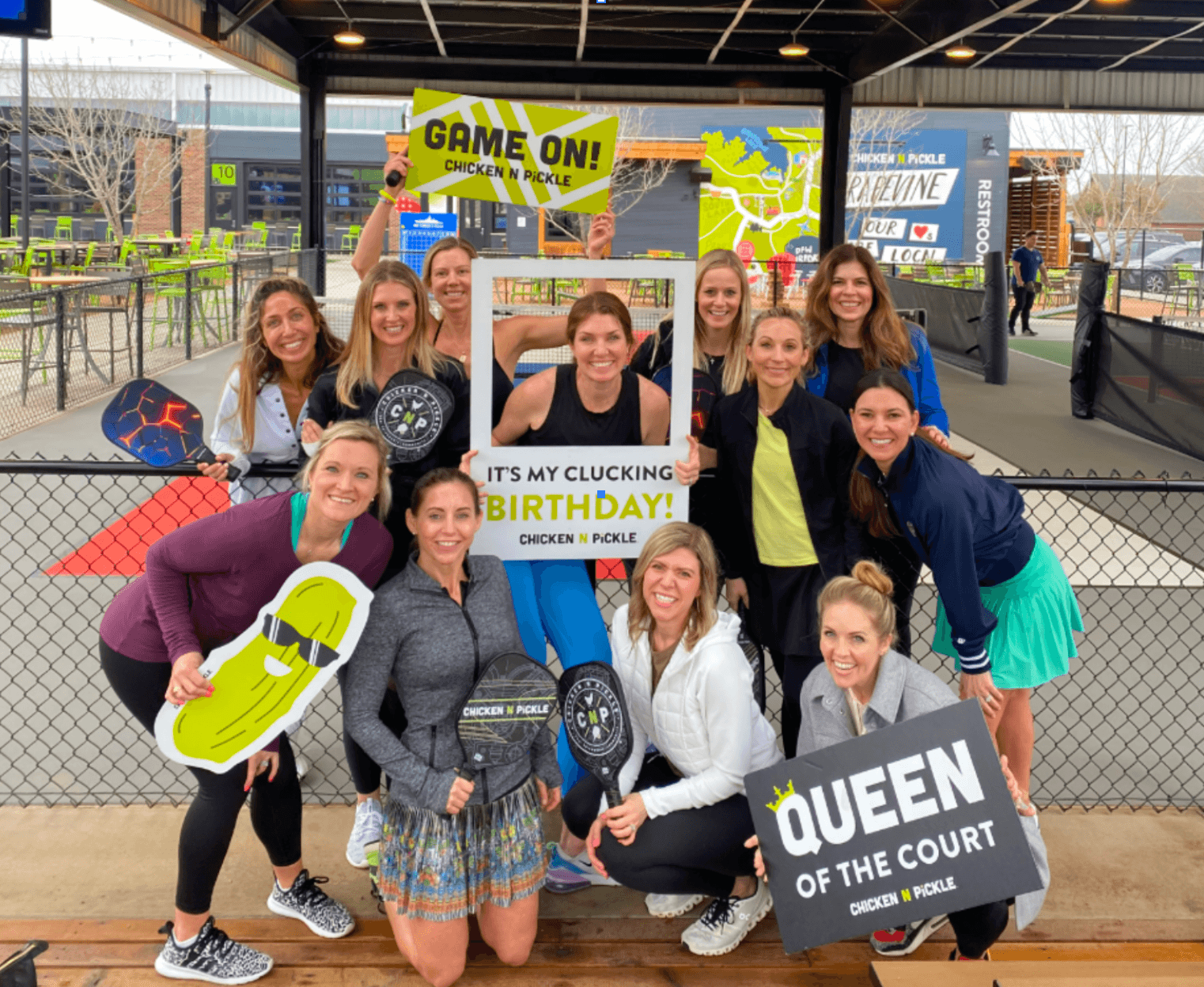 A group of women poses at a pickleball court, smiling and holding signs such as Game On!, Its My Clucking Birthday!, and Queen of the Court. Some hold paddles; the setting is outdoors under a covered area.