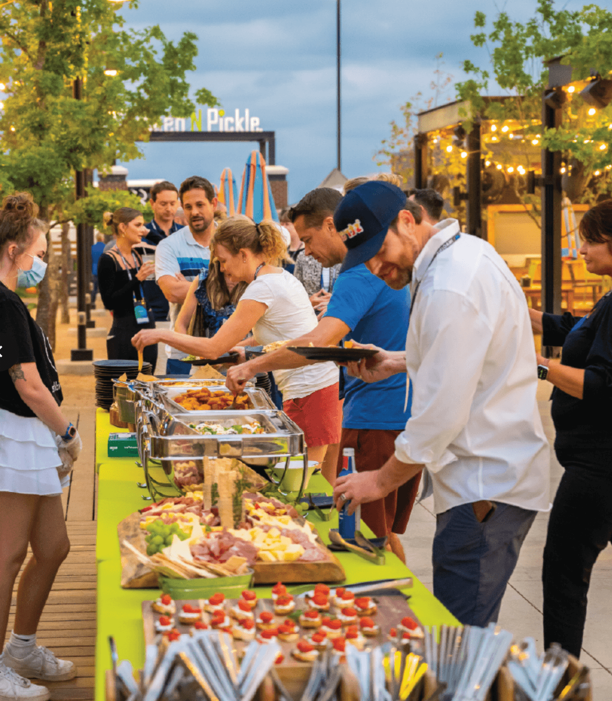 People serving themselves food from a buffet table outdoors, with various appetizers and snacks displayed. Others converse in the background under string lights, creating a lively, festive atmosphere. Trees and outdoor decor are visible.
