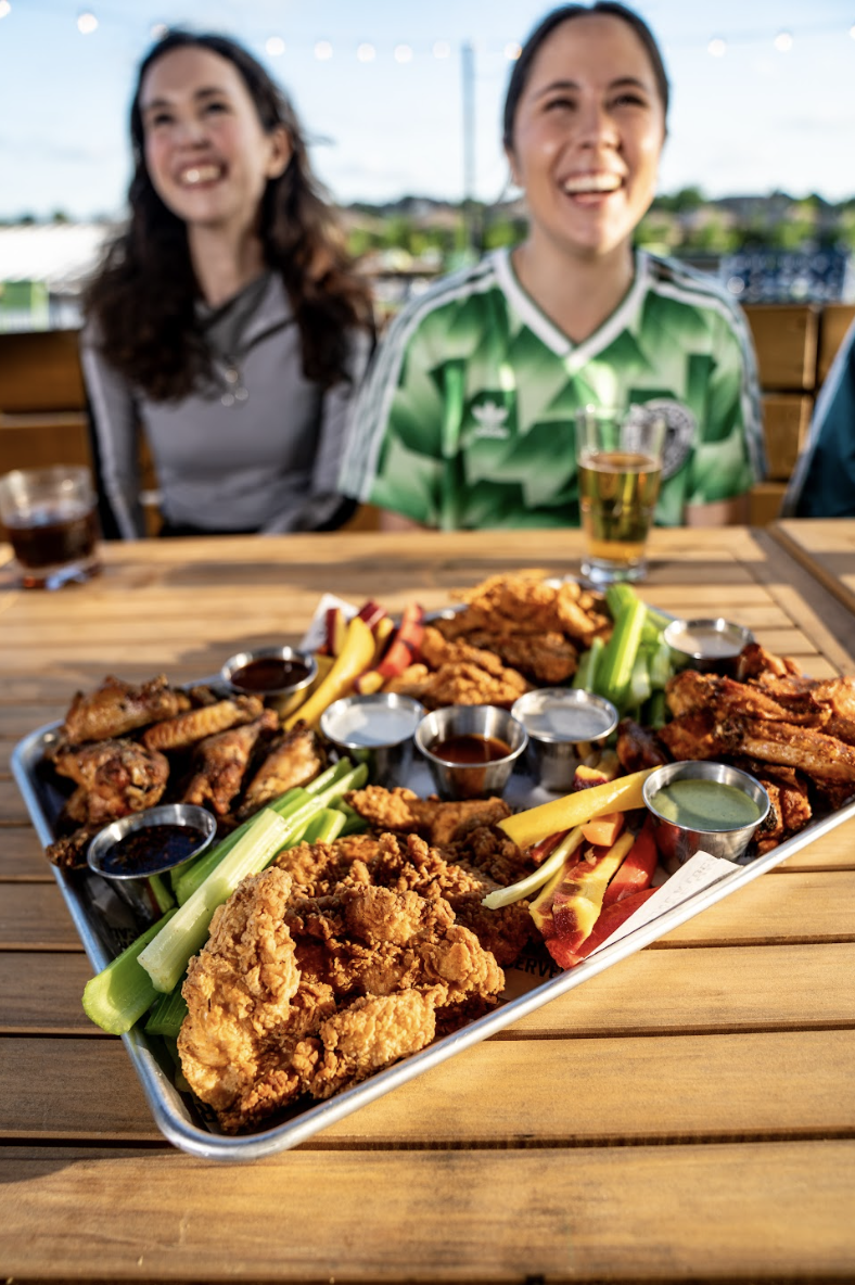 Two smiling women sit at an outdoor table with drinks, behind a tray of fried chicken, chicken wings, celery, and dipping sauces under bright daylight.