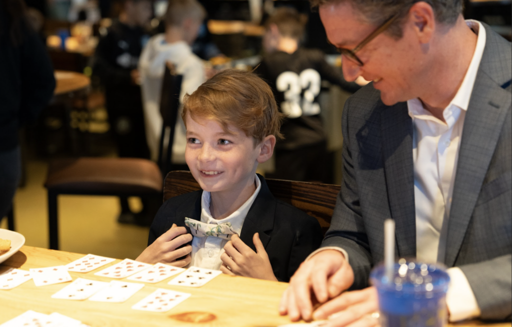 A smiling boy in a suit holds cash to his chest while sitting at a table with playing cards spread out, next to a man in glasses and a suit jacket, in a lively indoor setting.