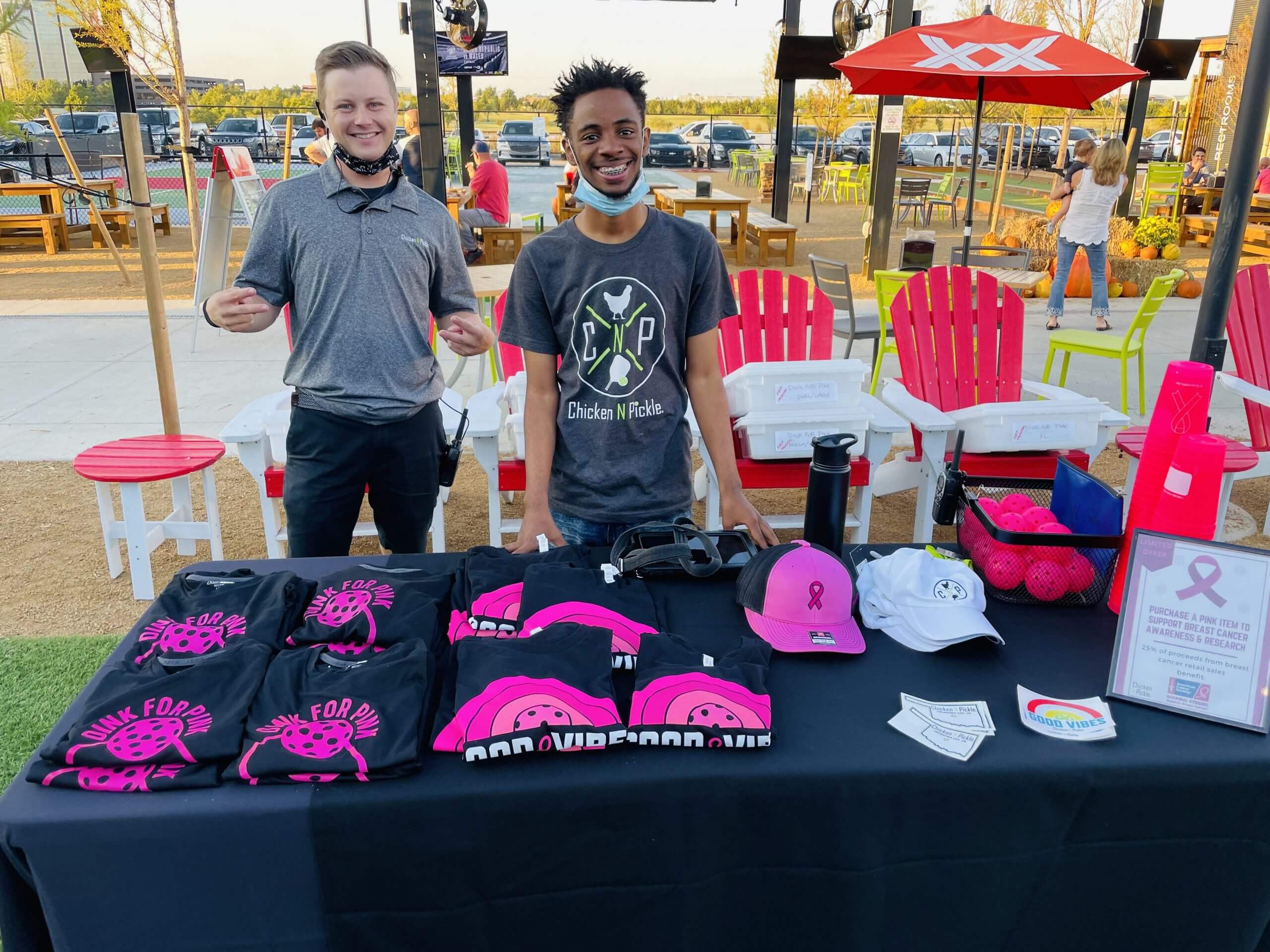 Two men stand smiling behind a table displaying black T-shirts, pink hats, a white cap, and pink promotional items at an outdoor event. There are chairs, tables, and people in the background.