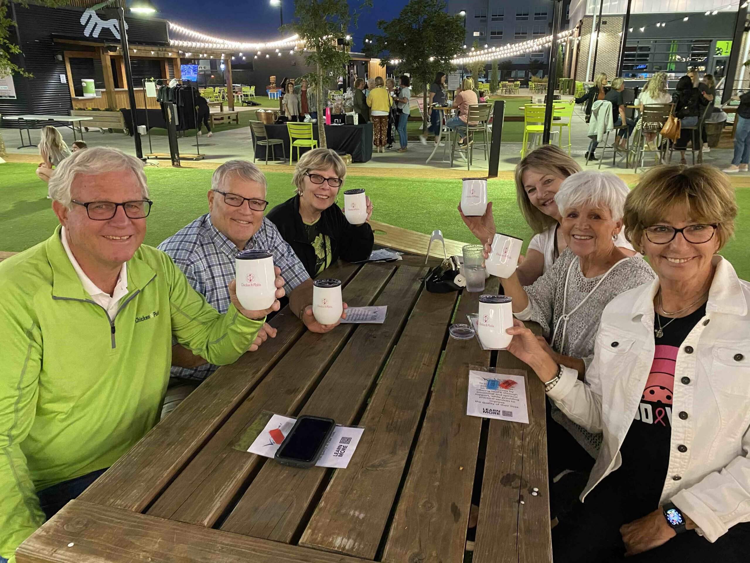 Six smiling older adults sit around a wooden picnic table outdoors in the evening, holding up white mugs. There are string lights overhead and people in the background. The atmosphere appears relaxed and cheerful.