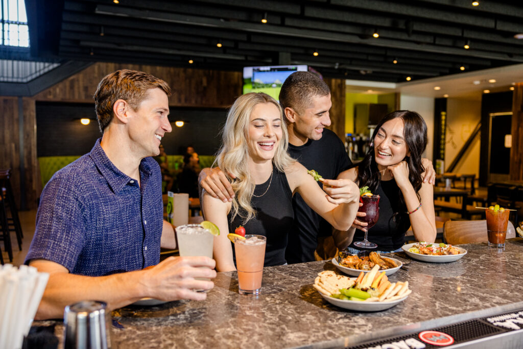 Four friends sit at a bar smiling and laughing, enjoying drinks and food. Plates of appetizers and colorful cocktails are on the counter in front of them. The setting is lively and casual.