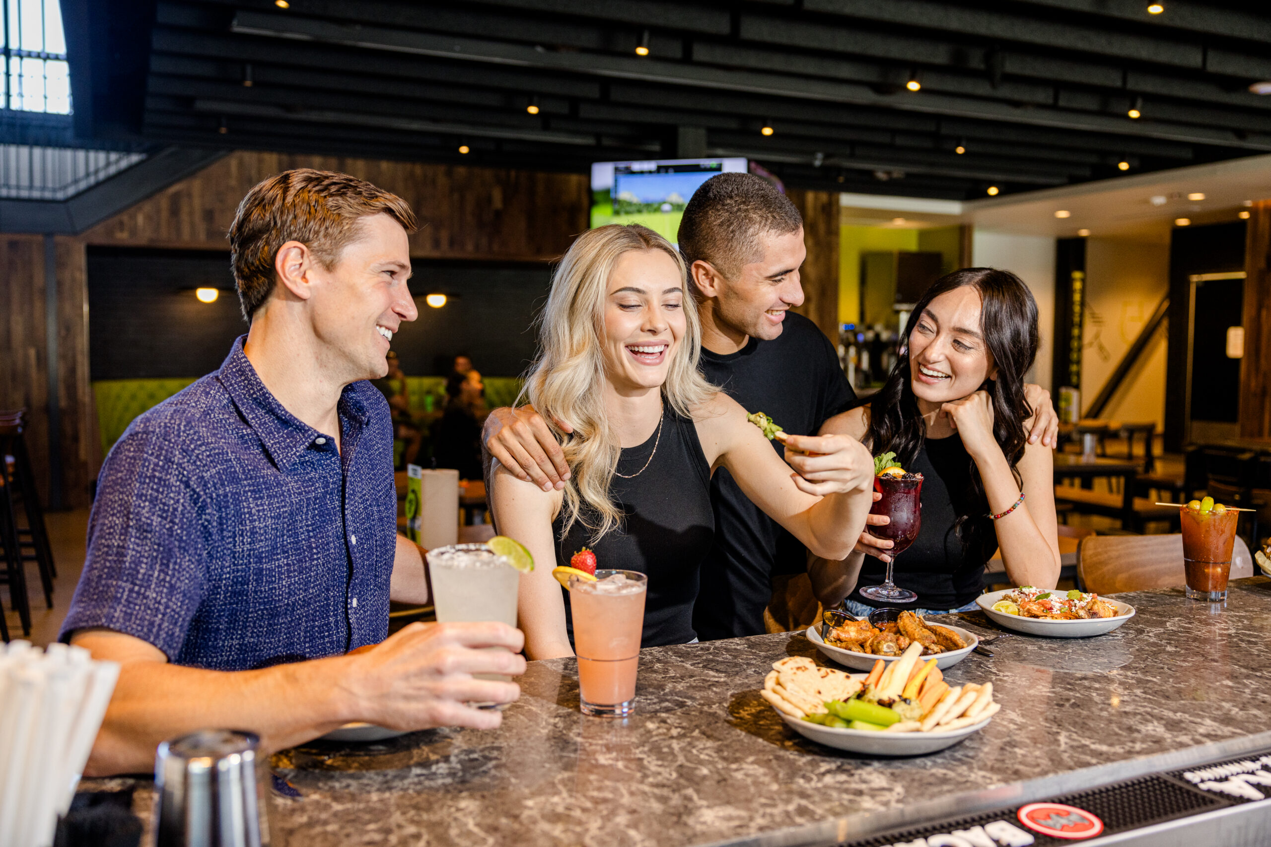 Four friends sit at a bar smiling and laughing, enjoying drinks and food. Plates of appetizers and colorful cocktails are on the counter in front of them. The setting is lively and casual.