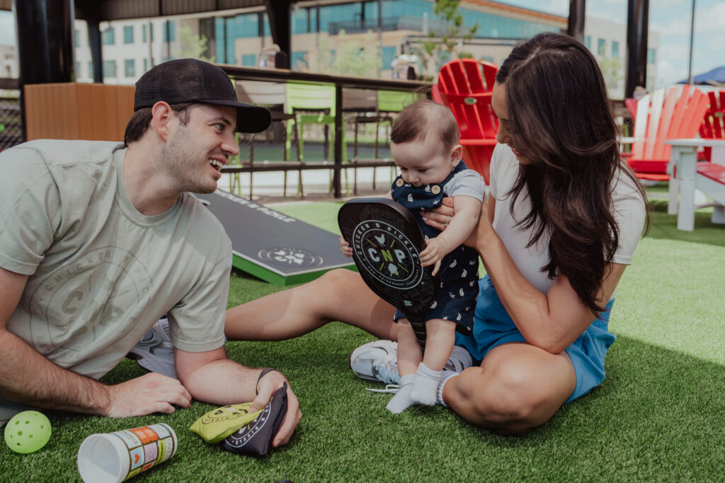 A man, woman, and baby sit on artificial grass outdoors at the 10% Giveback Night at Chicken N Pickle NKC. The man smiles at the baby holding a small paddle, while the woman helps steady the baby. Colorful chairs and bean bags brighten the background.