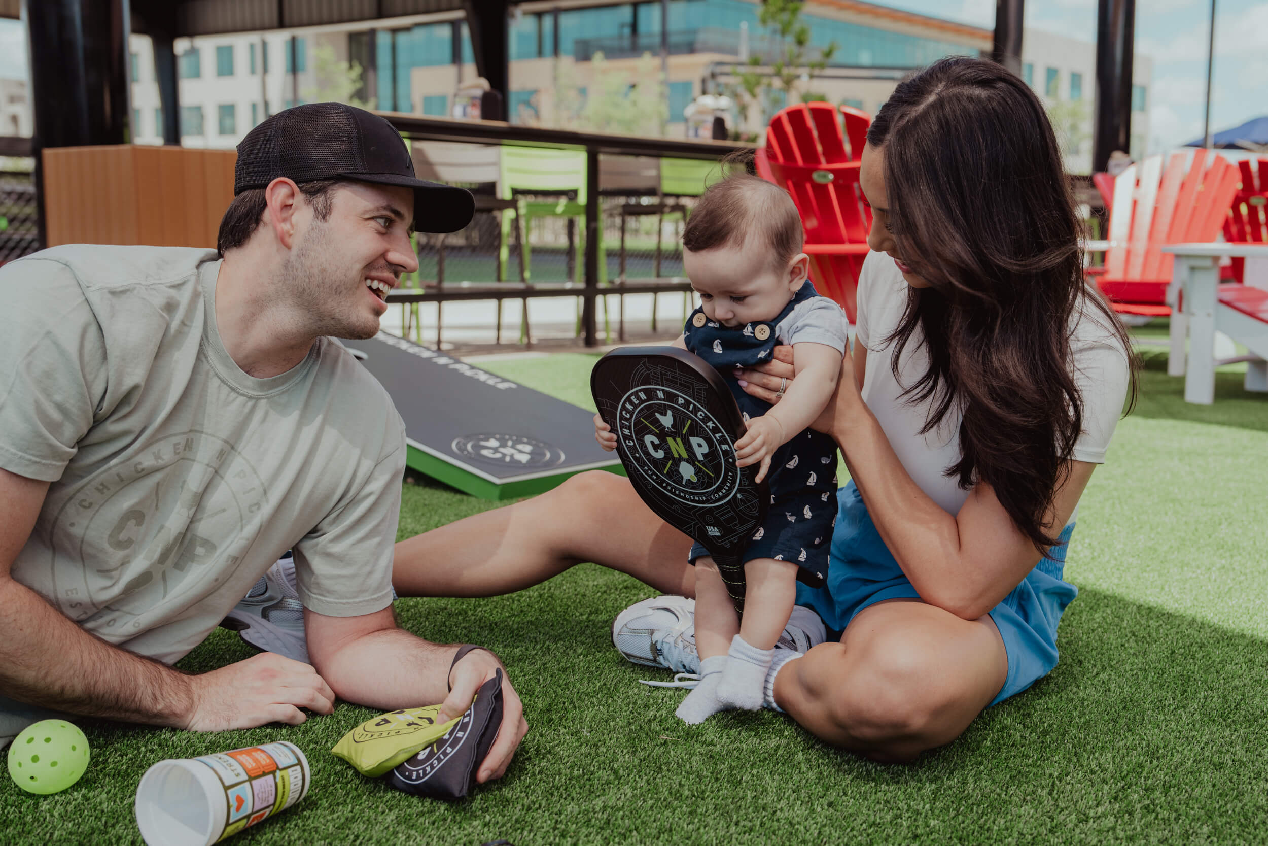 A man, woman, and baby sit on artificial grass outdoors at the 10% Giveback Night at Chicken N Pickle NKC. The man smiles at the baby holding a small paddle, while the woman helps steady the baby. Colorful chairs and bean bags brighten the background.