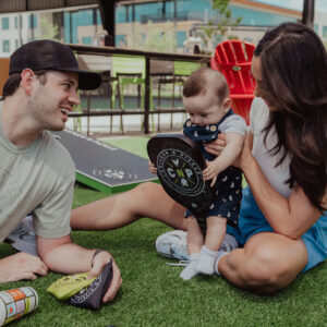A man, woman, and baby sit on artificial grass outdoors during 10% Giveback Night at Chicken N Pickle NKC. The man smiles at the baby, who holds a small paddle. Bright chairs and a cornhole board create a playful, relaxed scene in the background.