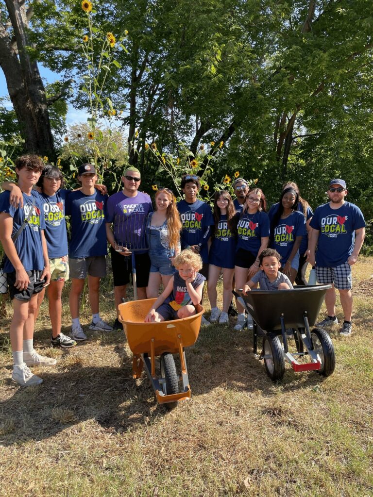 A group of people pose outdoors in front of tall sunflowers. Most wear matching blue OUR LOCAL shirts. Two children sit in wheelbarrows at the front, and trees fill the background. It is a sunny day.