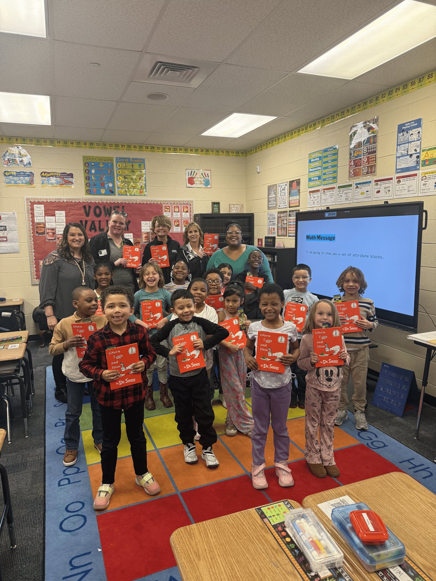 A group of young students in a classroom smile and hold up red books. Several adults stand behind them, also smiling. The classroom is colorful with educational posters and a large screen in the background.