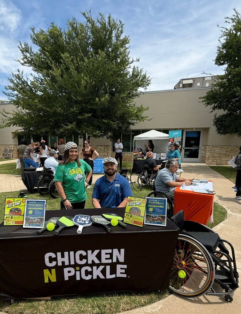 Two people, one standing and one sitting in a wheelchair, are behind a Chicken N Pickle table with pickleball paddles and flyers. Other people and tables are set up outdoors in front of a building. Trees and a sidewalk are visible.