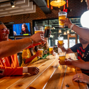 A group of people sit around a wooden table in a bar, smiling and raising glasses of beer and soda in a celebratory toast. A TV and large windows are visible in the background.