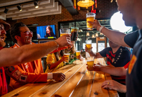 A group of people sit around a wooden table in a bar, smiling and raising glasses of beer and soda in a celebratory toast. A TV and large windows are visible in the background.