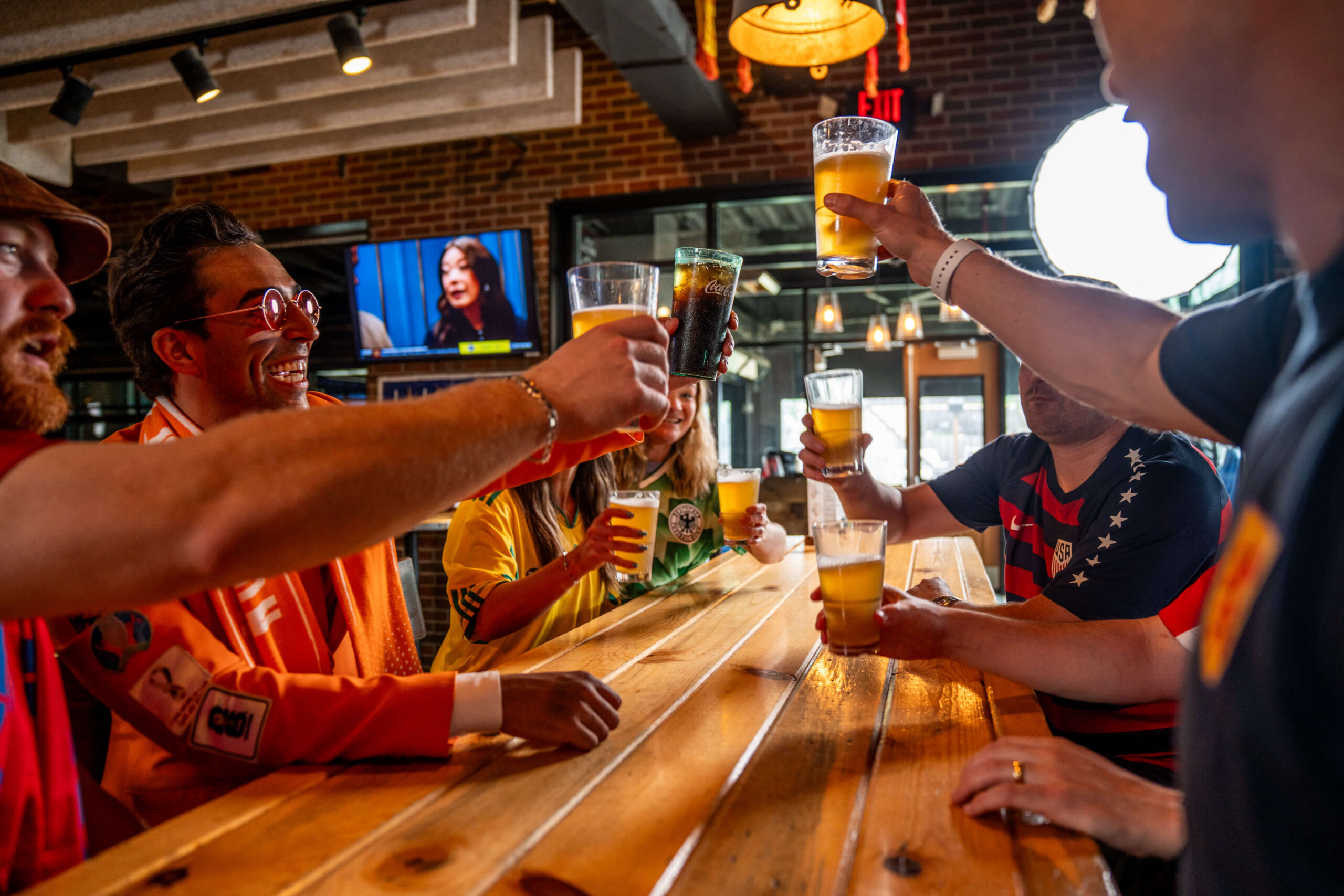 A group of people sit around a wooden table in a bar, smiling and raising glasses of beer and soda in a celebratory toast. A TV and large windows are visible in the background.