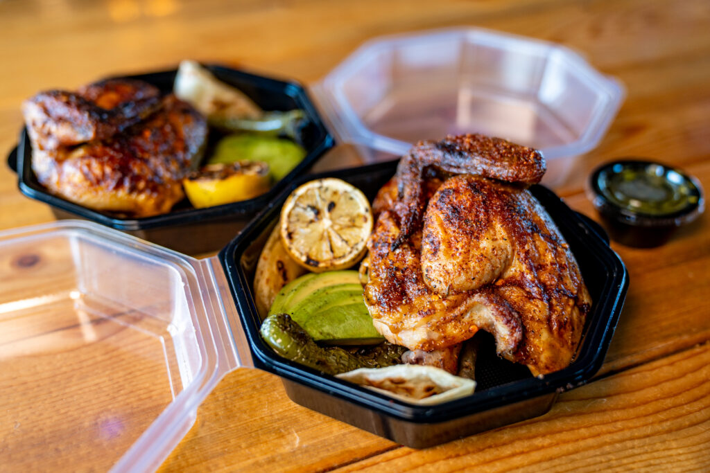 Two black takeout containers filled with grilled chicken, roasted vegetables, and lemon slices are placed on a wooden table. One container is open, and a small cup of sauce is nearby.