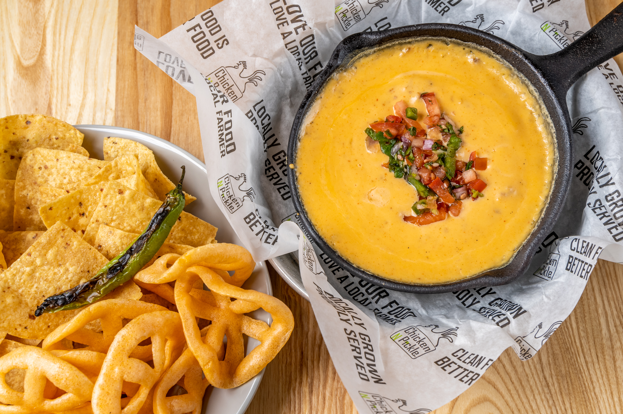 A skillet of creamy cheese dip topped with pico de gallo is served alongside tortilla chips, fried wagon wheel snacks, and a grilled chili pepper on a wooden table.