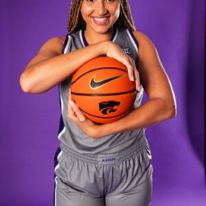 A female basketball player in a gray K-State uniform smiles and poses holding a basketball against a purple background.