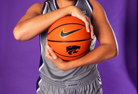 A female basketball player in a gray K-State uniform smiles and poses holding a basketball against a purple background.