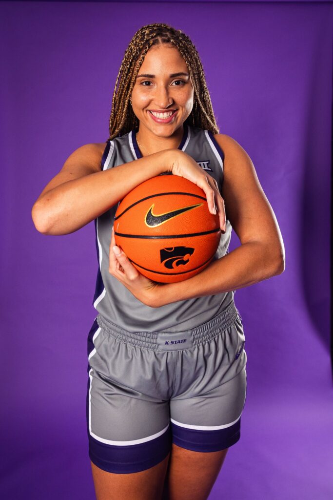 A female basketball player in a gray K-State uniform smiles and poses holding a basketball against a purple background.