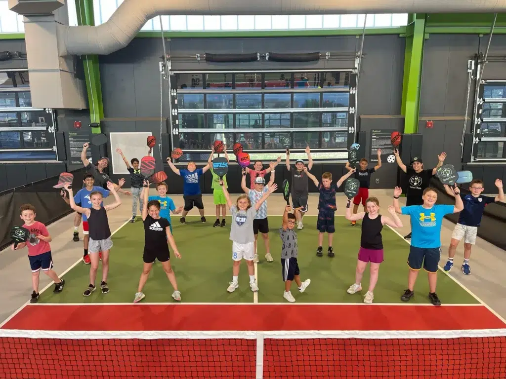A group of kids stands on an indoor tennis court, all smiling and raising one arm while holding tennis rackets. The court is brightly lit with green and red surfaces, and large windows are visible in the background.