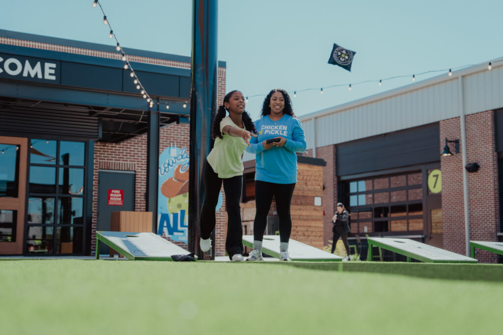Two women play cornhole outdoors on green turf near a building under string lights. One woman throws a bean bag as the other watches and smiles.