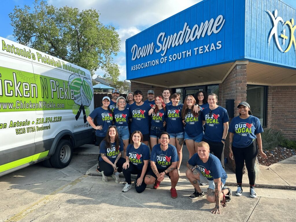 A group of people in matching Our NPickle shirts stands smiling outside the Down Syndrome Association of South Texas building, next to a Chicken N Pickle van.