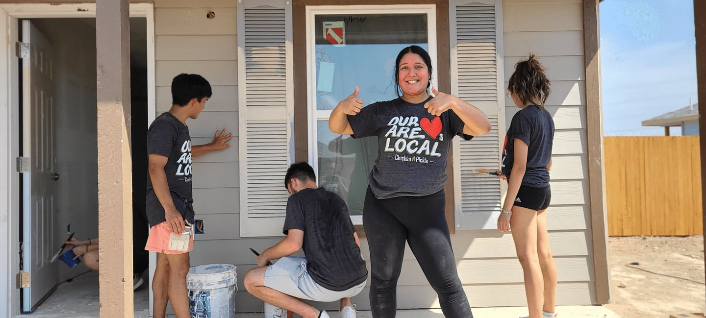 Four people paint the exterior of a house on a sunny day. One woman stands smiling and giving a thumbs up; all wear matching “Our Heart Are Local” shirts. Two paint the wall, one is crouched, and one faces away.