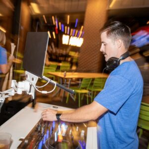 A DJ wearing headphones and a blue shirt mixes music at an indoor venue with green chairs and blurred colorful lights in the background.