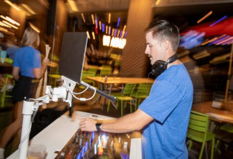 A DJ wearing headphones and a blue shirt mixes music at an indoor venue with green chairs and blurred colorful lights in the background.