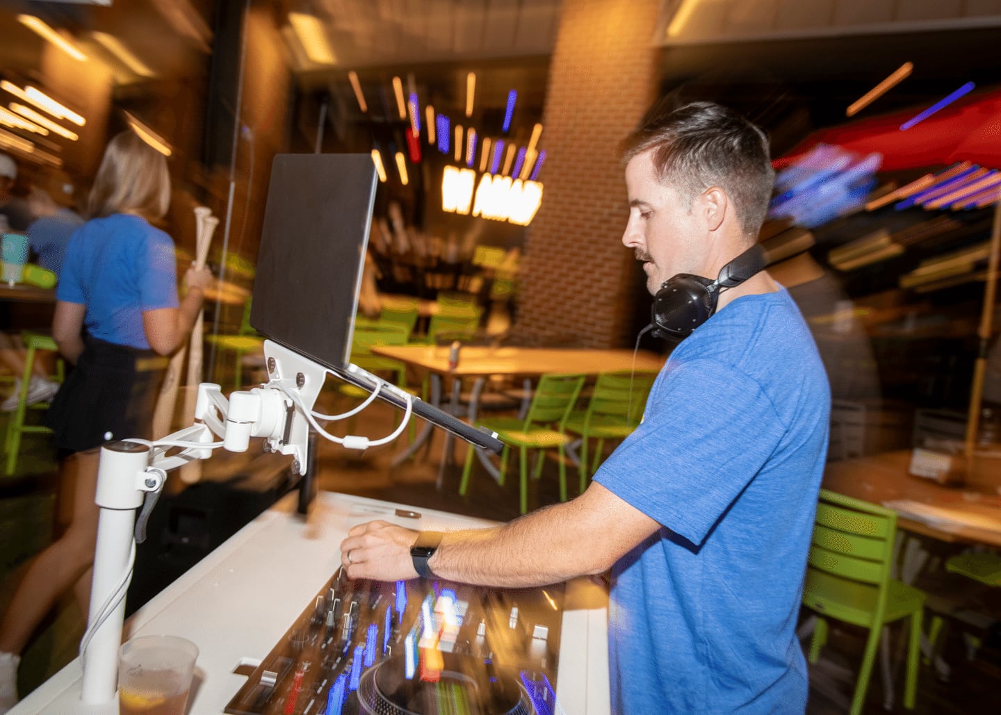A DJ wearing headphones and a blue shirt mixes music at an indoor venue with green chairs and blurred colorful lights in the background.