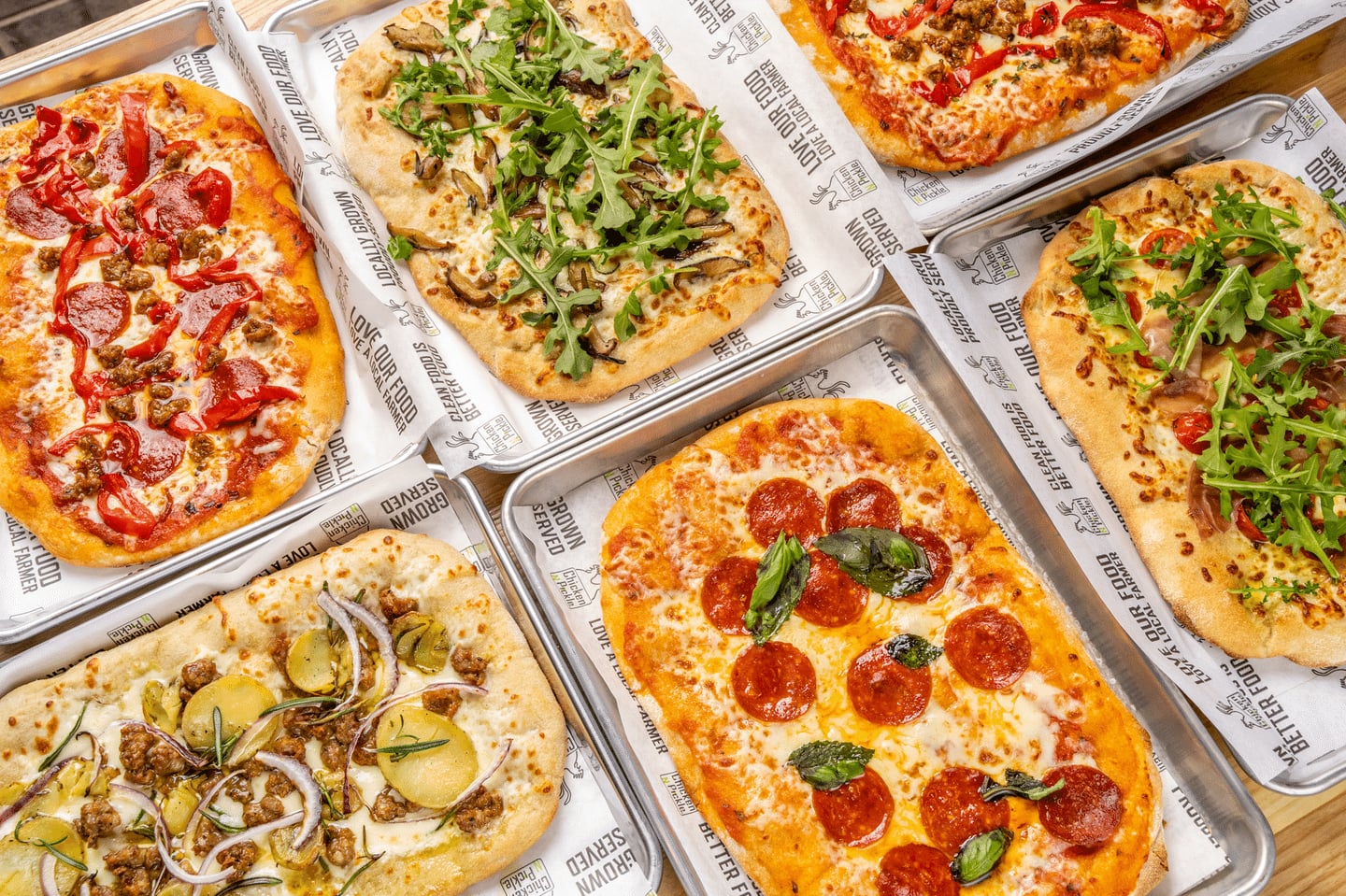 Six rectangular pizzas on metal trays, each topped with different ingredients like pepperoni, arugula, onions, red peppers, and mushrooms, are arranged on branded parchment paper. The pizzas feature a variety of colorful toppings.