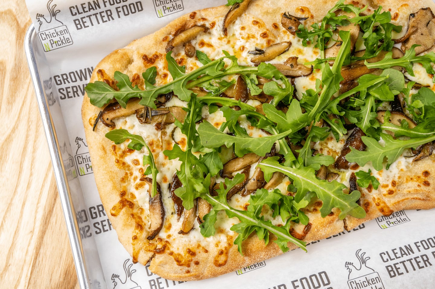 A rectangular flatbread topped with melted cheese, mushrooms, and fresh arugula, served on branded paper in a metal tray.